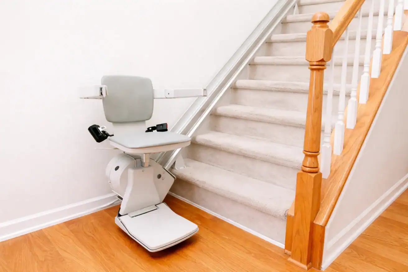 A stair lift chair is installed at the base of a carpeted staircase with wooden handrails in a residential setting.