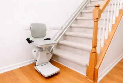 A stair lift chair is installed at the base of a carpeted staircase with wooden handrails in a residential setting.
