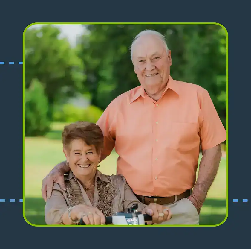 An elderly man and woman pose outdoors; the man stands with his hand on the woman's shoulder while she sits on a mobility scooter. Both are smiling. Trees and grass are in the background.