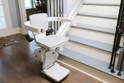 A stair lift chair is installed along the side of a staircase, with the seat positioned at the bottom ready for use.