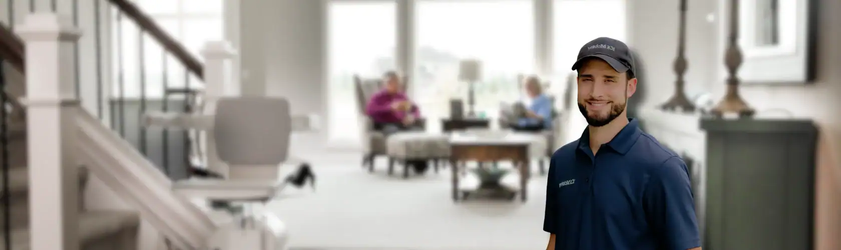 A technician in a uniform stands in the foreground of a bright living room with two people sitting and talking in the background.