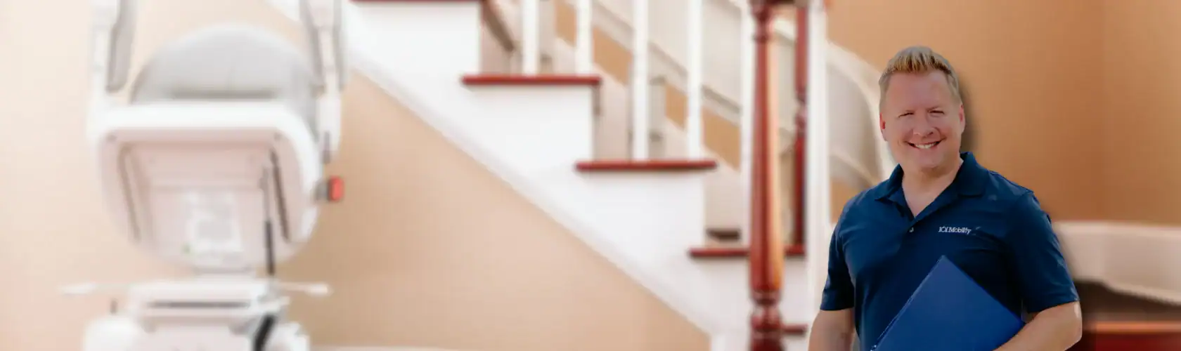 A smiling man in a blue polo shirt stands holding a folder in front of a staircase with a stairlift installed on the side.