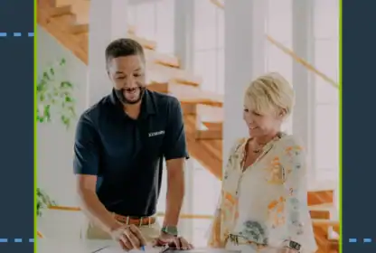 A man in a navy shirt points at a document on a counter while a woman in a patterned blouse watches and smiles; they stand in a modern, well-lit interior.