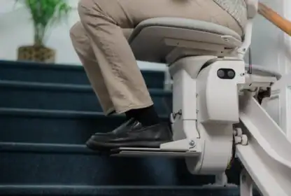 A person seated on a stair lift rides up a carpeted staircase, with one hand resting on the armrest and feet on the footrest.