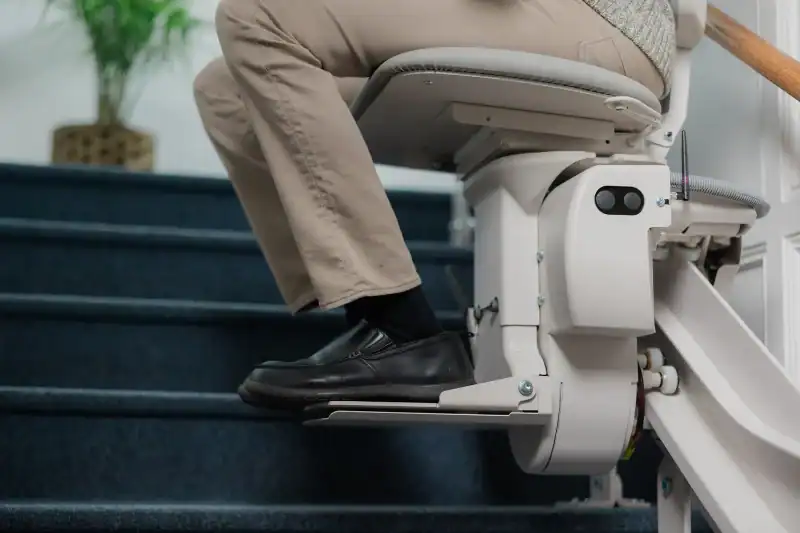 A person seated on a stair lift rides up a carpeted staircase, with one hand resting on the armrest and feet on the footrest.