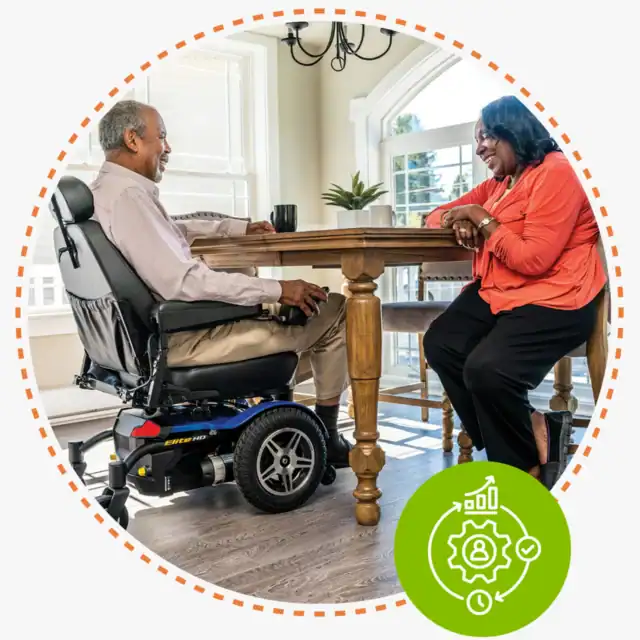 An older man in a power wheelchair and an older woman sit across from each other at a wooden dining table, smiling and talking in a sunlit room.