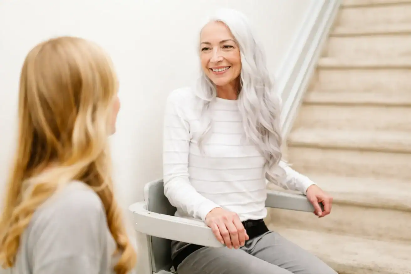 An older woman with long gray hair sits on a stair lift, smiling at a younger woman with blonde hair standing nearby.