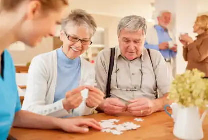 Two older adults and a caregiver assemble a jigsaw puzzle at a table, while another elderly couple converses in the background of a bright room.