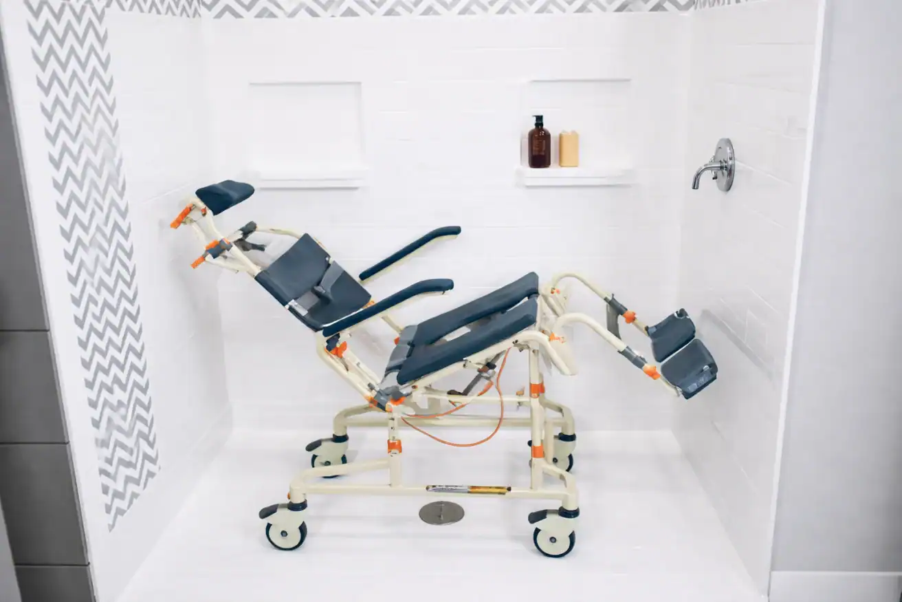 A reclining shower chair with wheels and adjustable leg and arm supports is positioned inside a tiled shower area.