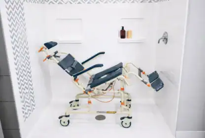 A reclining shower chair with wheels and adjustable leg and arm supports is positioned inside a tiled shower area.