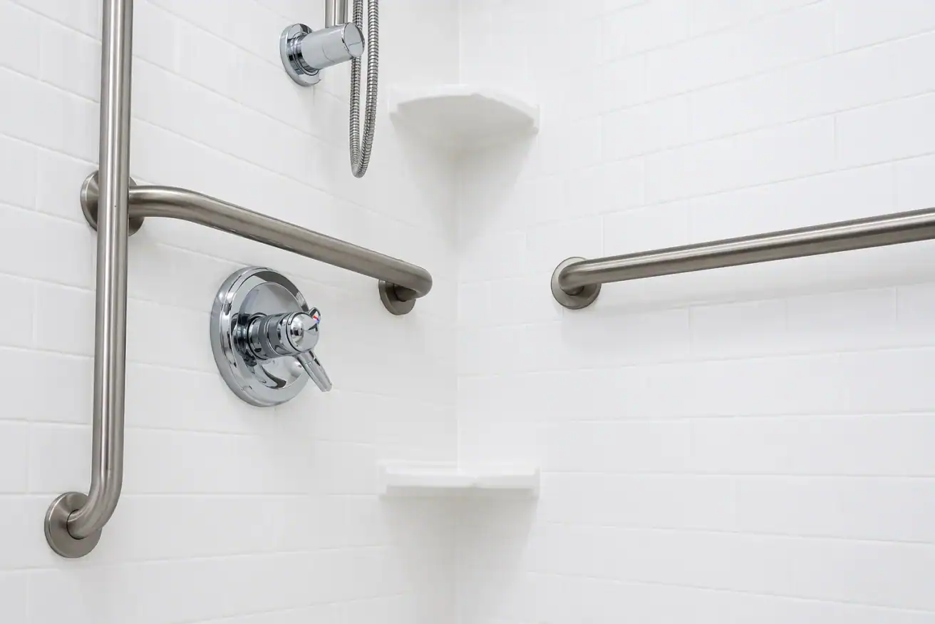 White tiled shower corner with metal grab bars, a round shower control, a handheld showerhead holder, and two corner shelves.