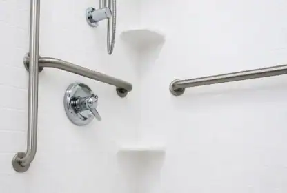 White tiled shower corner with metal grab bars, a round shower control, a handheld showerhead holder, and two corner shelves.