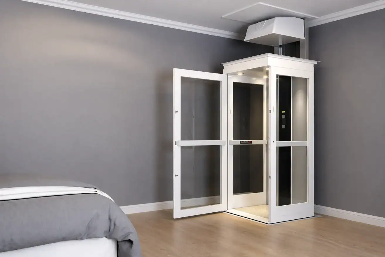 A small residential elevator with glass doors is installed in a modern room with gray walls and wood flooring. The elevator door is open, and part of a bed is visible in the foreground.