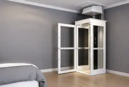 A small residential elevator with glass doors is installed in a modern room with gray walls and wood flooring. The elevator door is open, and part of a bed is visible in the foreground.