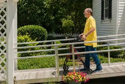 An older man in a yellow shirt uses a walker to move up a wooden ramp outside a white house with greenery and flowers nearby.