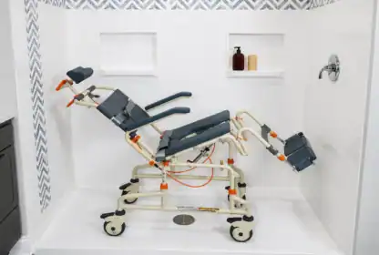 A reclining, wheeled shower chair with padded supports is positioned inside a white tiled shower with a wall-mounted faucet and toiletries on built-in shelves.