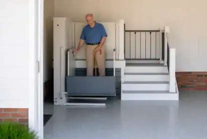 An older man with a cane stands on a wheelchair lift next to a staircase in a residential garage.