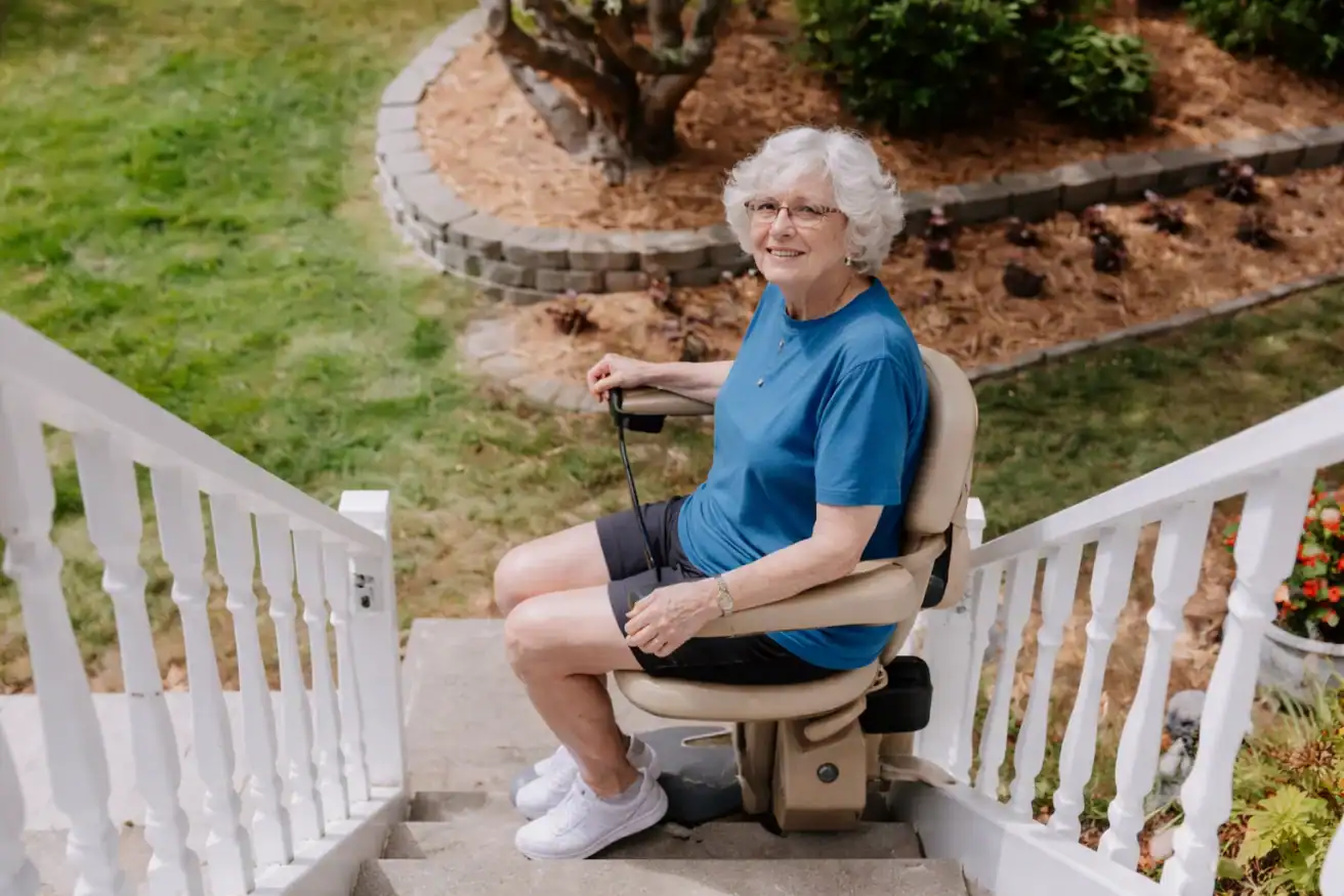 An older woman in a blue shirt sits on a motorized stair lift outside, ascending a set of outdoor stairs with a garden in the background.