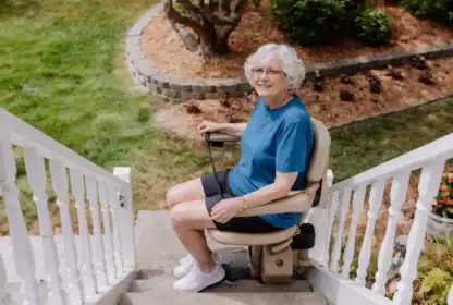 An older woman in a blue shirt sits on a motorized stair lift outside, ascending a set of outdoor stairs with a garden in the background.