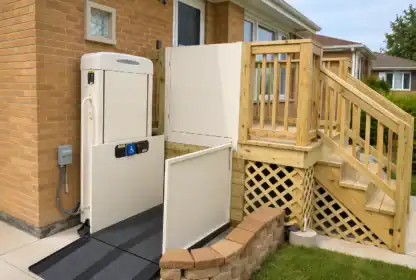 A residential wheelchair lift is installed next to a wooden porch and stairs, providing accessible entry to the house.