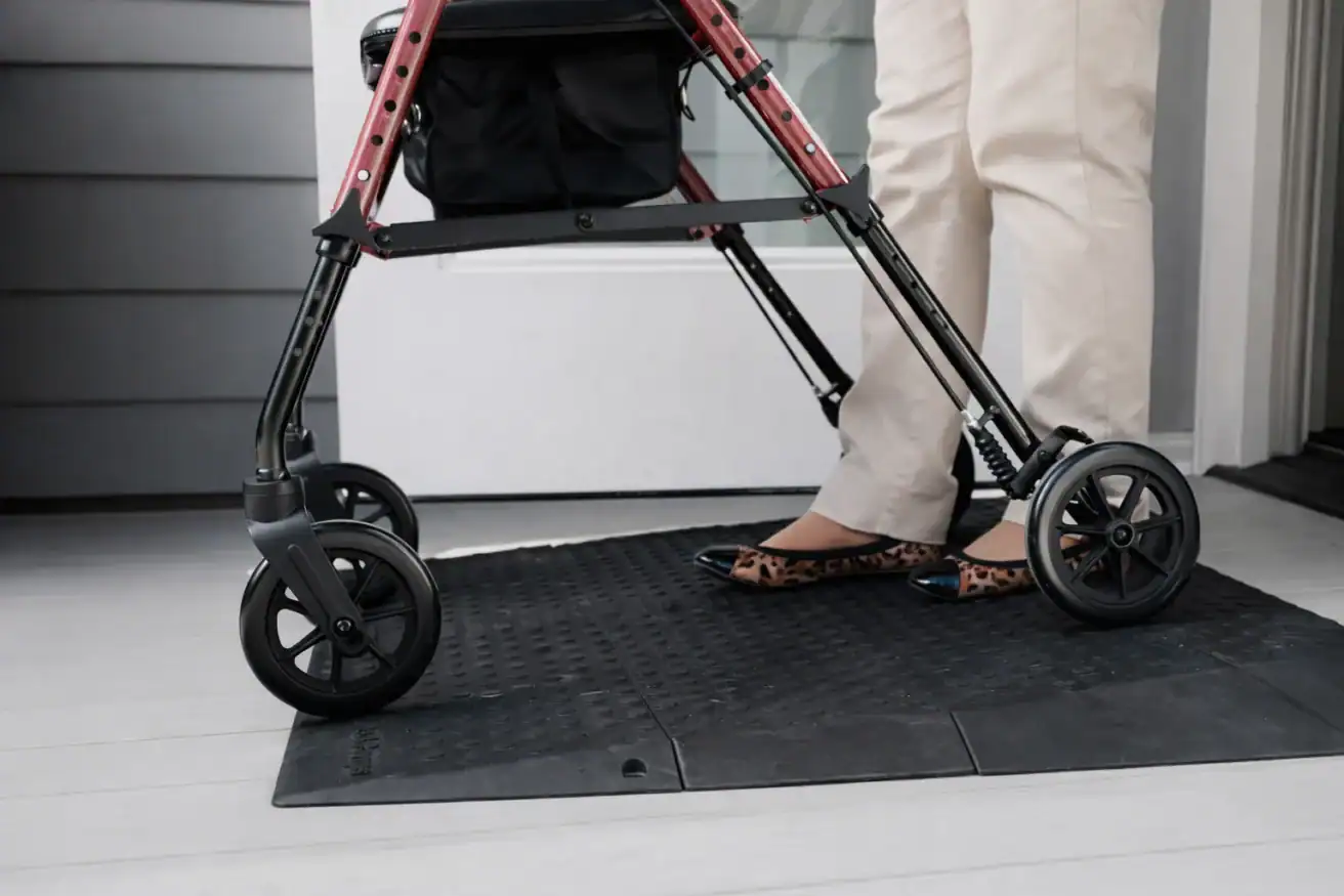 A person wearing beige pants and leopard print shoes stands behind a red walker on a rubber mat in front of a door.