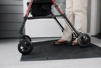 A person wearing beige pants and leopard print shoes stands behind a red walker on a rubber mat in front of a door.