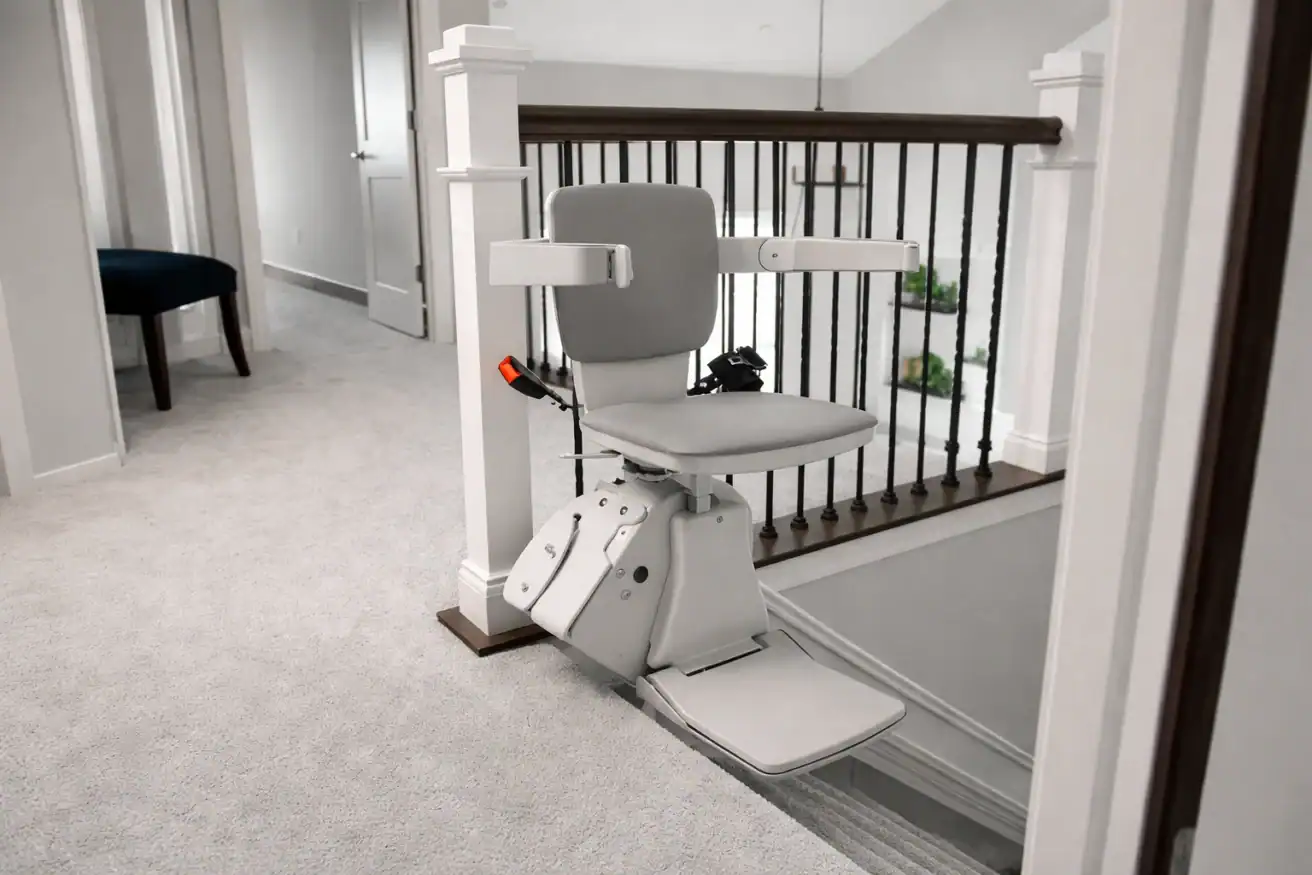 A motorized stair lift with a gray seat and armrests is installed at the top of a carpeted staircase in a residential home.