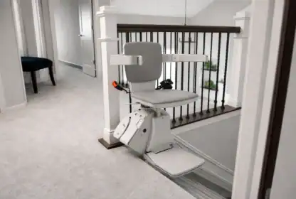 A motorized stair lift with a gray seat and armrests is installed at the top of a carpeted staircase in a residential home.