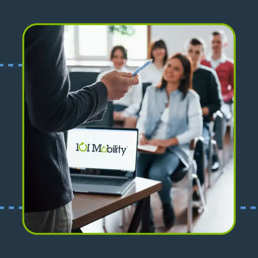 A person gives a presentation to a seated group in a classroom setting; a laptop displaying the 101 Mobility logo is on the desk in front.