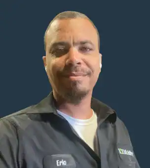 A man wearing a dark work shirt with a name tag that reads "Eric" stands against a plain dark background, smiling at the camera.