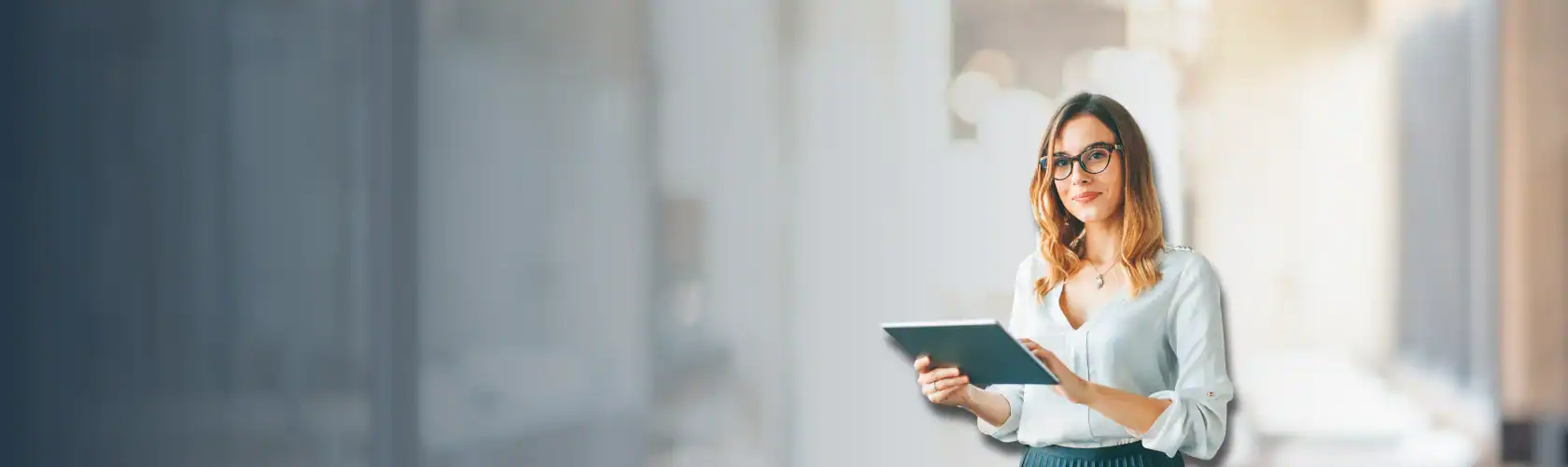 A woman wearing glasses and business attire holds a tablet while standing in a modern, well-lit office environment.