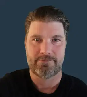 A man with short, slicked-back hair and a beard wearing a black shirt poses in front of a plain dark blue background.