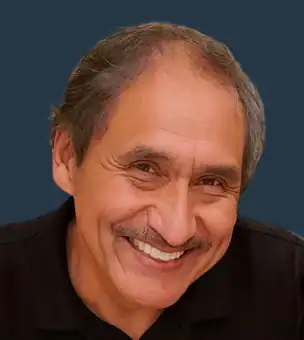 A middle-aged man with a mustache and salt-and-pepper hair smiles, wearing a black collared shirt against a plain dark blue background.
