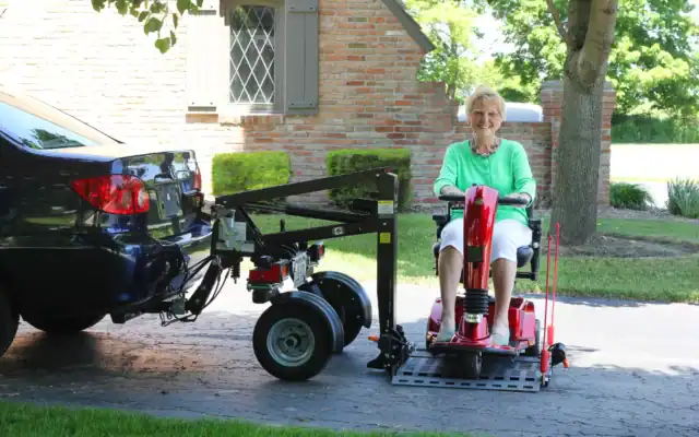 An older woman sits on a red mobility scooter beside a blue car equipped with a mechanical lift in a residential driveway.