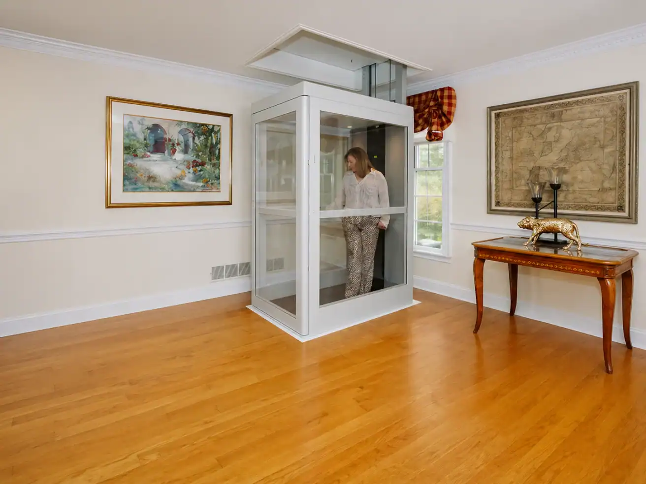 A person stands inside a glass residential elevator in a spacious room with hardwood floors, artwork on the walls, and a decorative table.