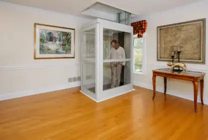 A person stands inside a glass residential elevator in a spacious room with hardwood floors, artwork on the walls, and a decorative table.