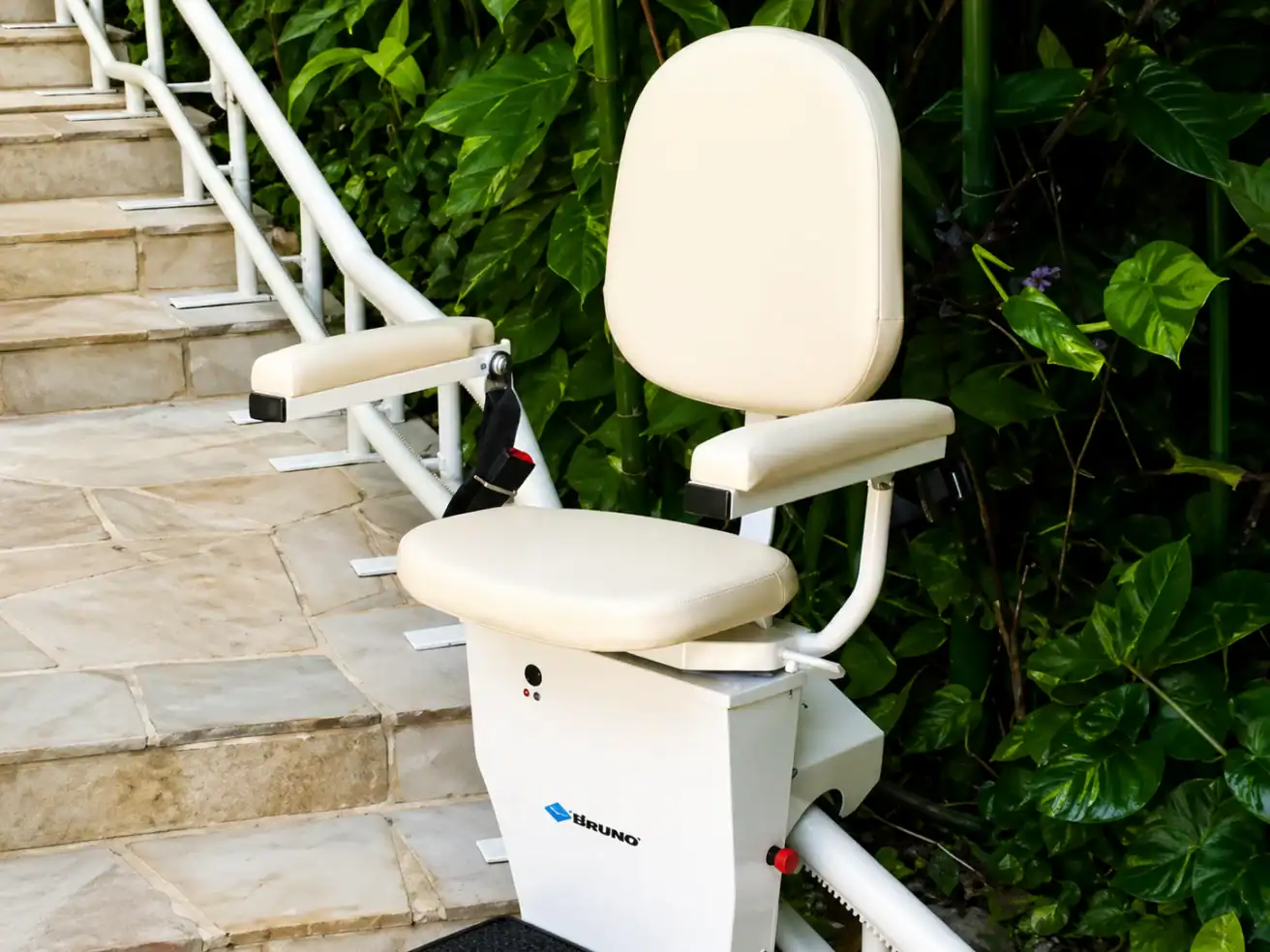 A stair lift with a padded seat and armrests is installed alongside an outdoor staircase next to green plants.