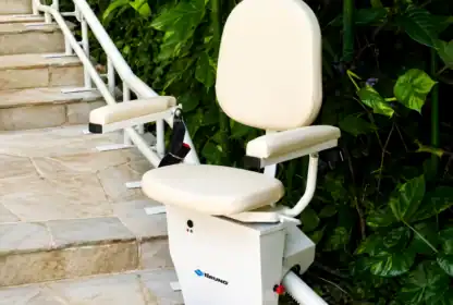 A stair lift with a padded seat and armrests is installed alongside an outdoor staircase next to green plants.