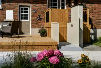 A residential outdoor lift beside a wooden deck provides wheelchair accessibility to the house; pink and yellow flowers grow in the foreground.