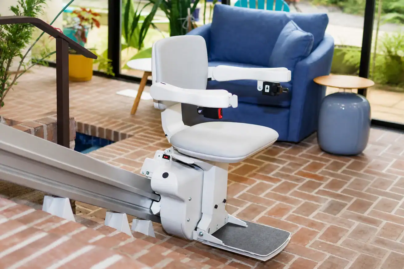 A stairlift with a padded seat and armrests is installed along indoor brick steps near a blue armchair and plants.