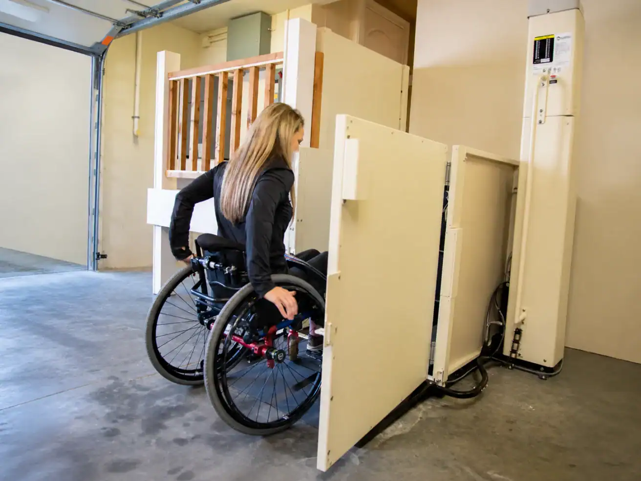 A woman in a wheelchair uses a vertical platform lift in a garage or indoor setting.