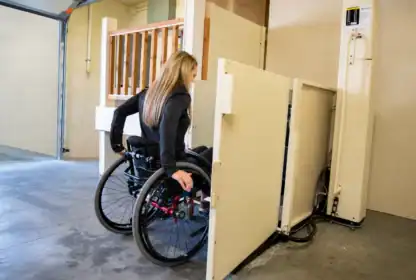 A woman in a wheelchair uses a vertical platform lift in a garage or indoor setting.