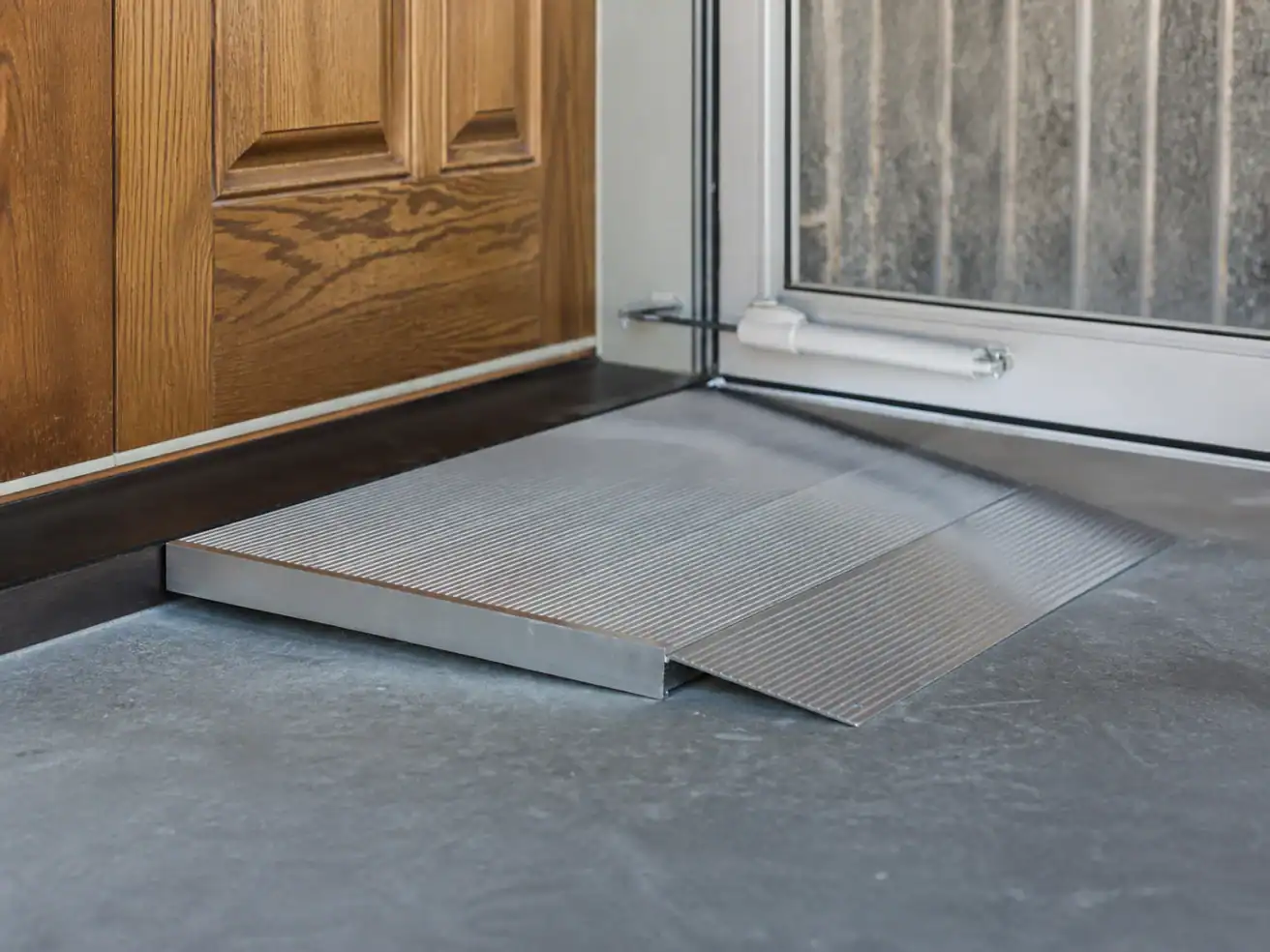 A metal wheelchair ramp is placed at the threshold between a wooden door and a glass door on a concrete floor.