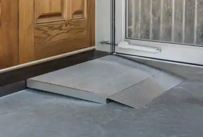 A metal wheelchair ramp is placed at the threshold between a wooden door and a glass door on a concrete floor.