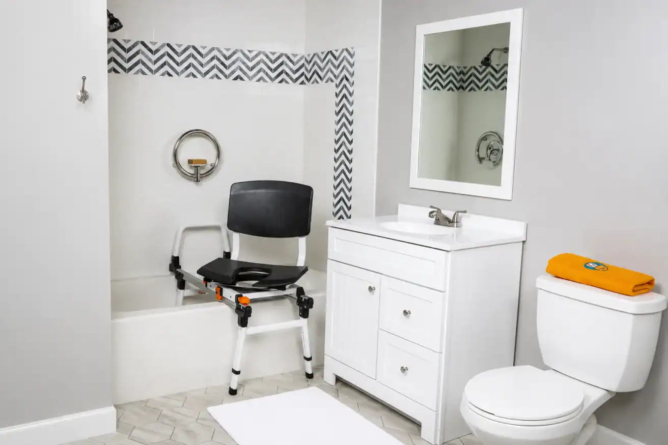 Accessible bathroom with a shower chair partially placed in a bathtub, a grab bar on the wall, a white vanity with a mirror, and a toilet with an orange towel on the tank.