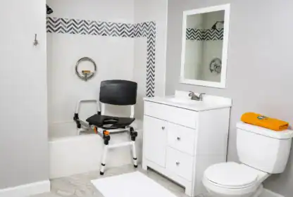 Accessible bathroom with a shower chair partially placed in a bathtub, a grab bar on the wall, a white vanity with a mirror, and a toilet with an orange towel on the tank.