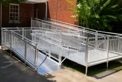 A metal accessibility ramp with railings provides wheelchair access to a brick building, surrounded by trees and concrete pavement.