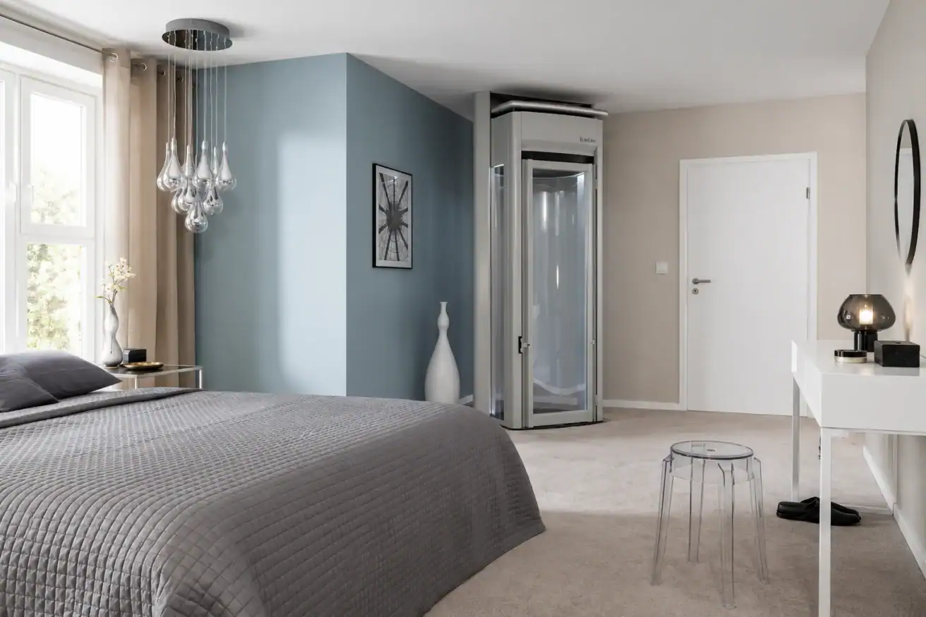 Modern bedroom with gray bedding, a small glass home elevator in the corner, a white desk, a clear stool, and decorative items against neutral walls.