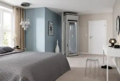 Modern bedroom with gray bedding, a small glass home elevator in the corner, a white desk, a clear stool, and decorative items against neutral walls.