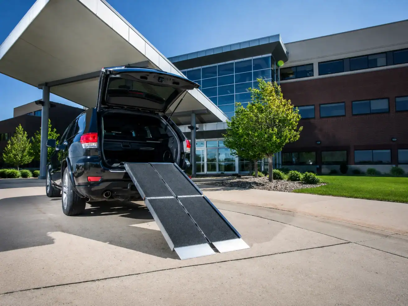 A black SUV with its trunk open and a wheelchair ramp extended onto a sidewalk outside a modern building.